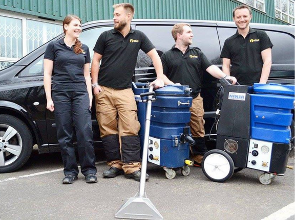 This is a photo of Chigwell Carpet Cleaning carpet cleaners (three men and one woman) standing in fromt of their black van, with two steam cleaning carpet machines next to them